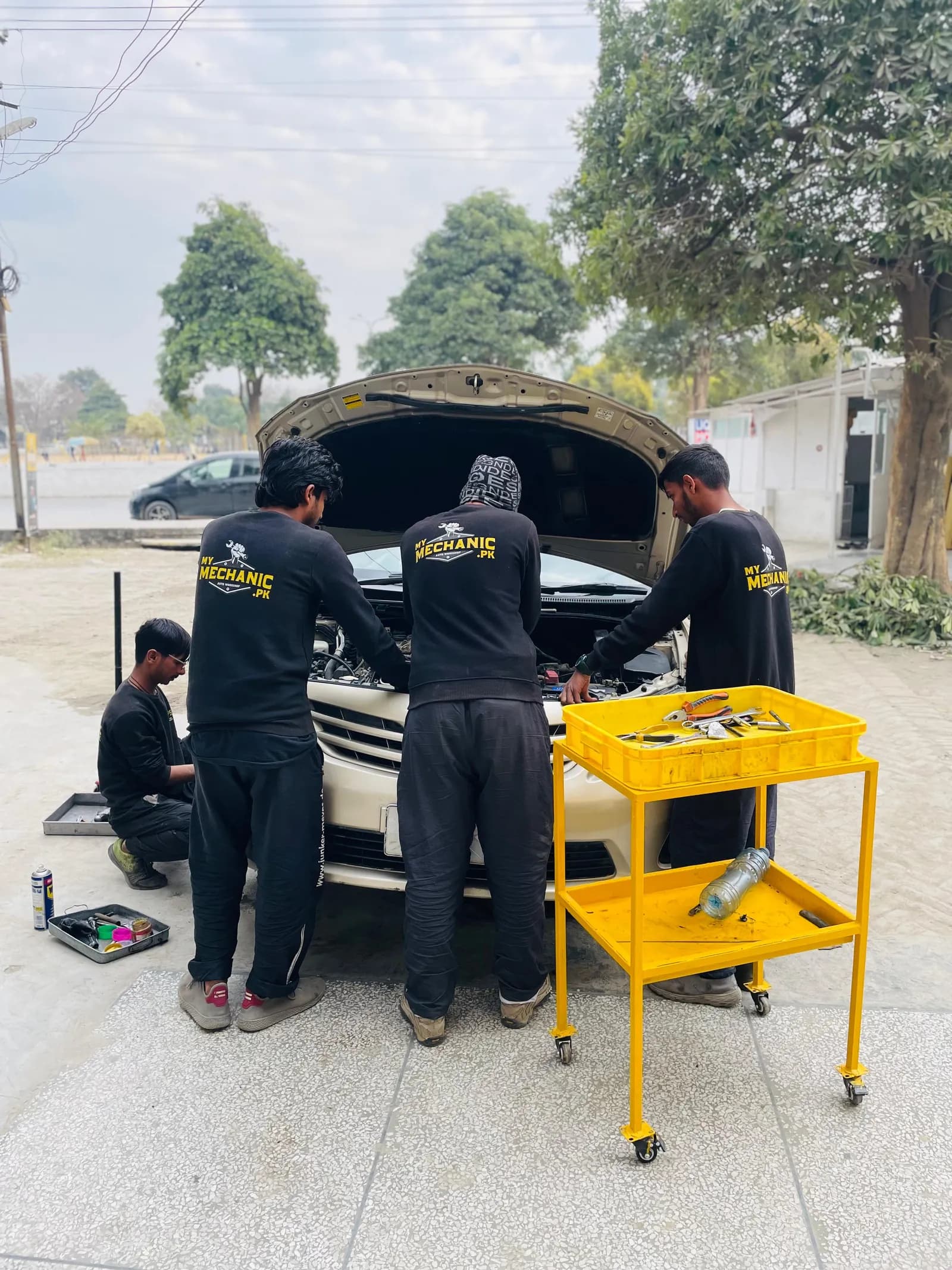Four My Mechanic technicians in uniform working on a car with a yellow tool trolley
